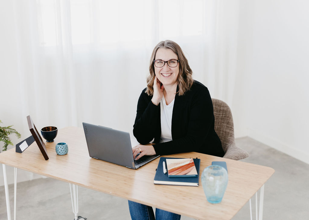 A woman sitting at a desk with a hand on a laptop keyboard and the other hand on the side of her neck. She is smiling at the camera and is wearing glasses, a white shirt and black sweater.